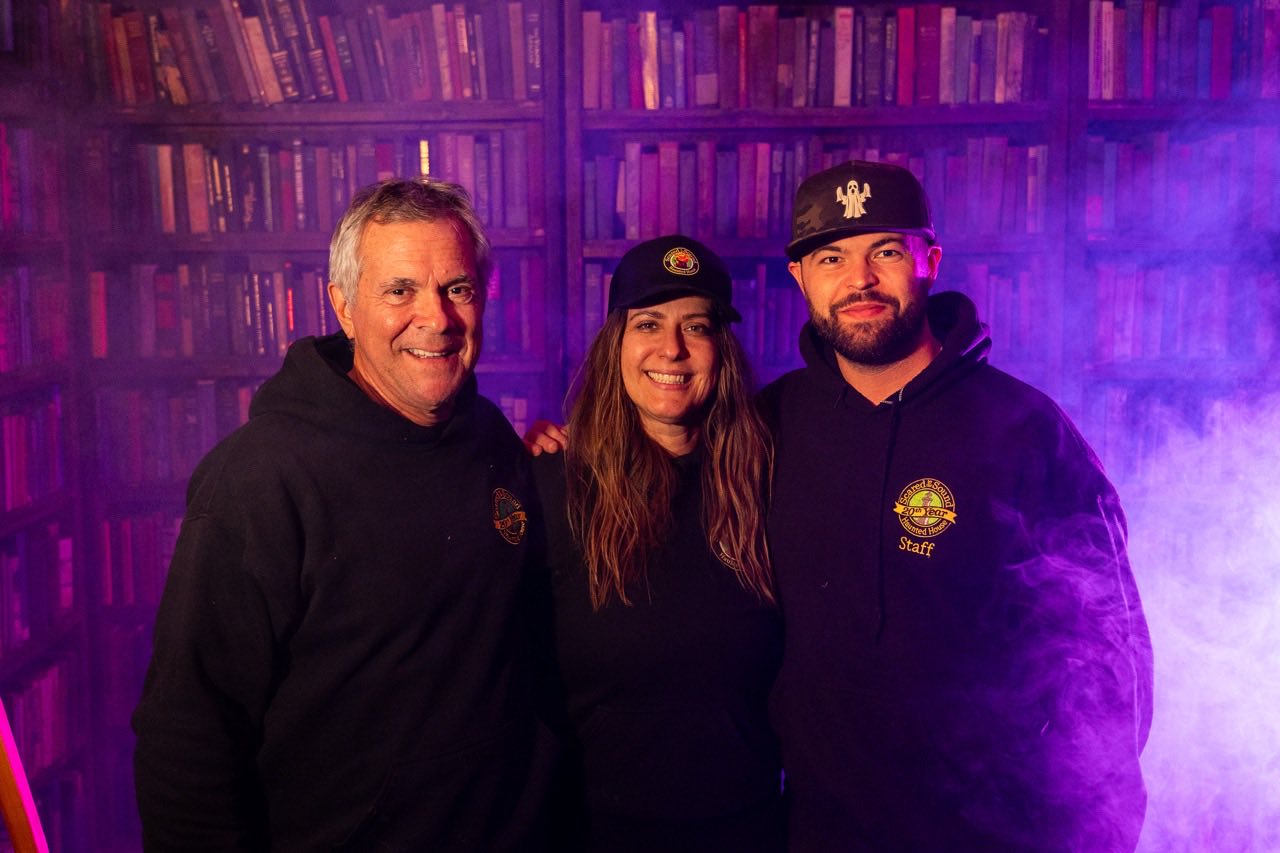 Three members of the Scared by the Sound team in staff hoodies, standing together in front of the haunt&rsquo;s library set under purple light and fog.